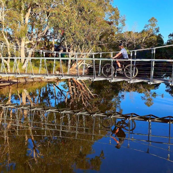 Mountain bike riders crossing a suspension bridge mirrored in a lake, Noosa Hinterland multi-day bike journey